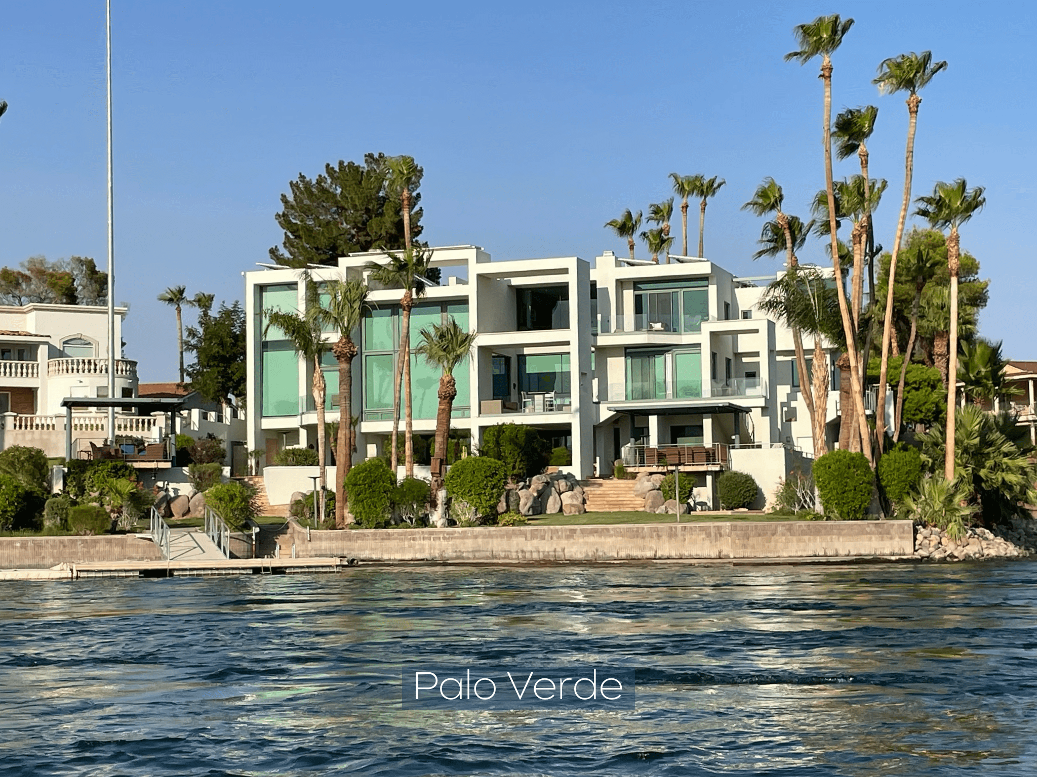 Modern white building with large windows by a waterfront and palm trees.